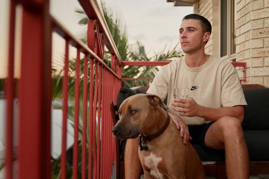 A young man on his balcony patting his dog and holding a glass of water. 