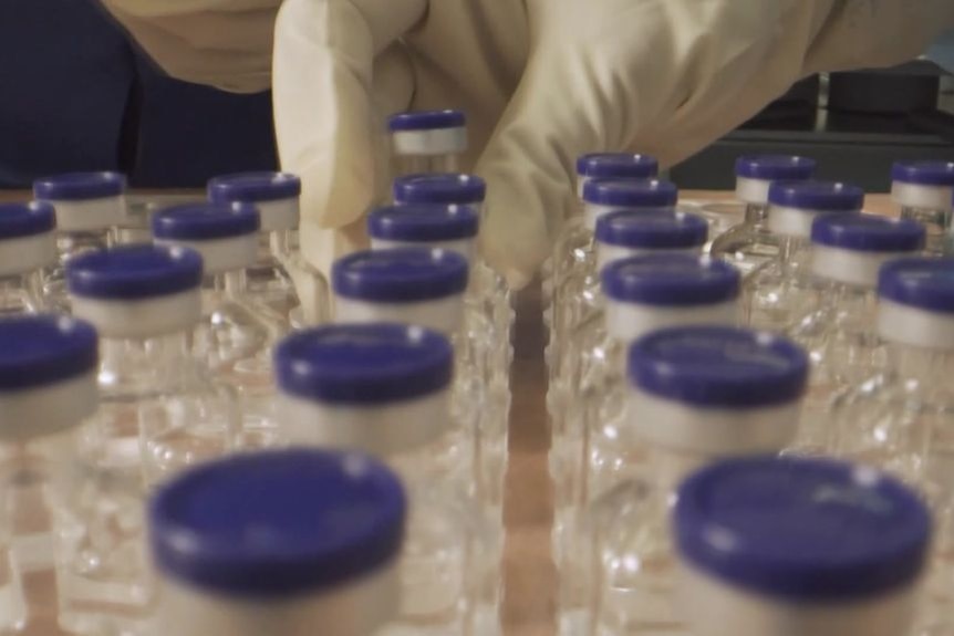 A close-up photo of a nurse's gloved hand reaching for COVID-19 vaccine vials 