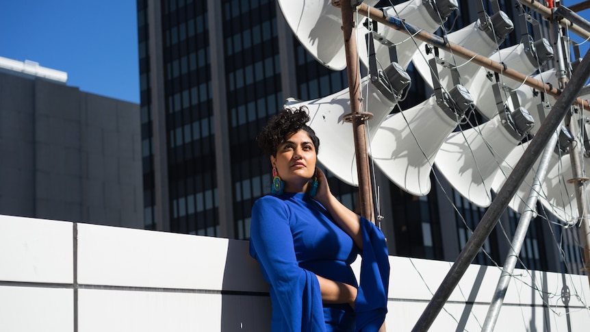 Singer Tara Tiba stands on building top beside  bank of speakers, wearing bright blue mini dress, with skyscrapers in background