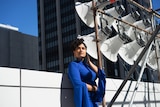 Singer Tara Tiba stands on building top beside  bank of speakers, wearing bright blue mini dress, with skyscrapers in background