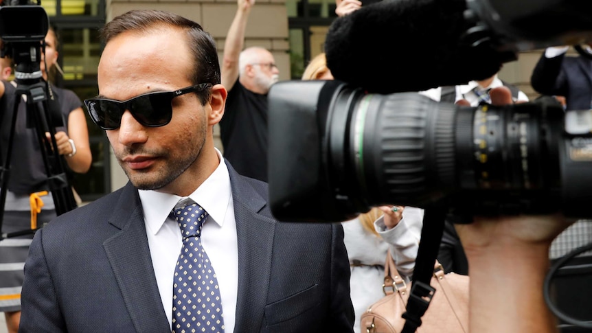 A man wearing sunglasses and a tailored suit walks through a media pack