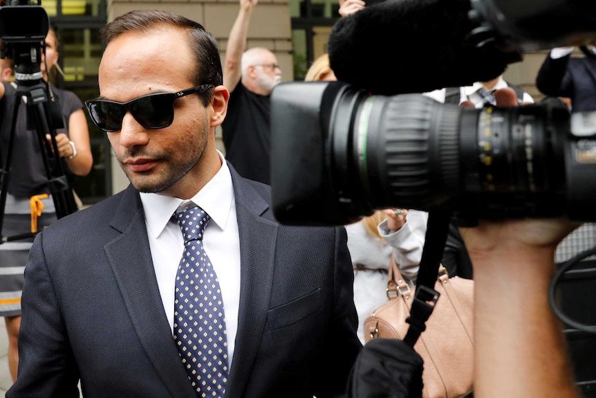 A man wearing sunglasses and a tailored suit walks through a media pack