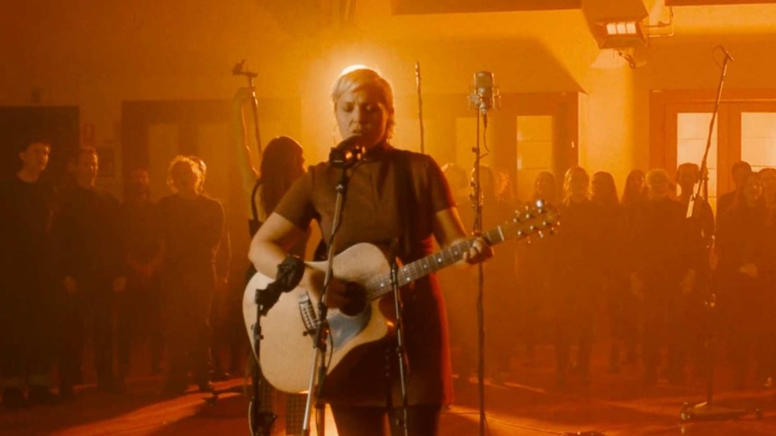 Liz is performing in a room lighted in red and orange with a choir in the background