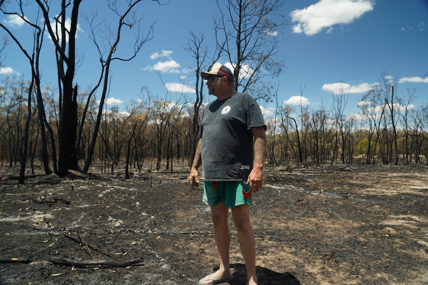a man wearing a cap and thongs stands in a blackened clearing in front of trees that have been burned out by a bushfire