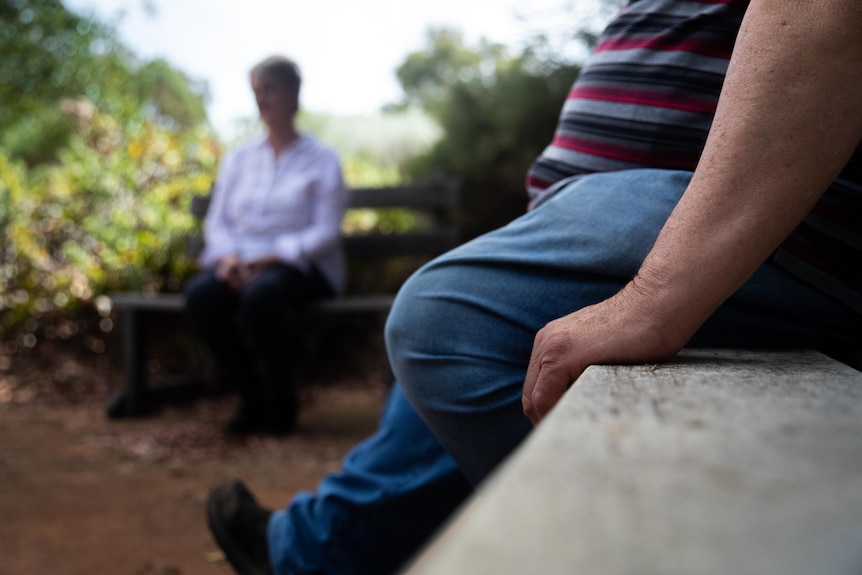 A man and a woman sitting on benches perpendicular to each other in a park.