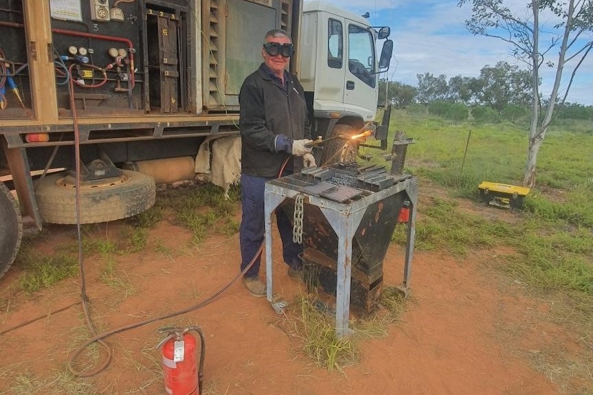 A man in goggles and metal working gear works at a welding table with a big grin on his face.