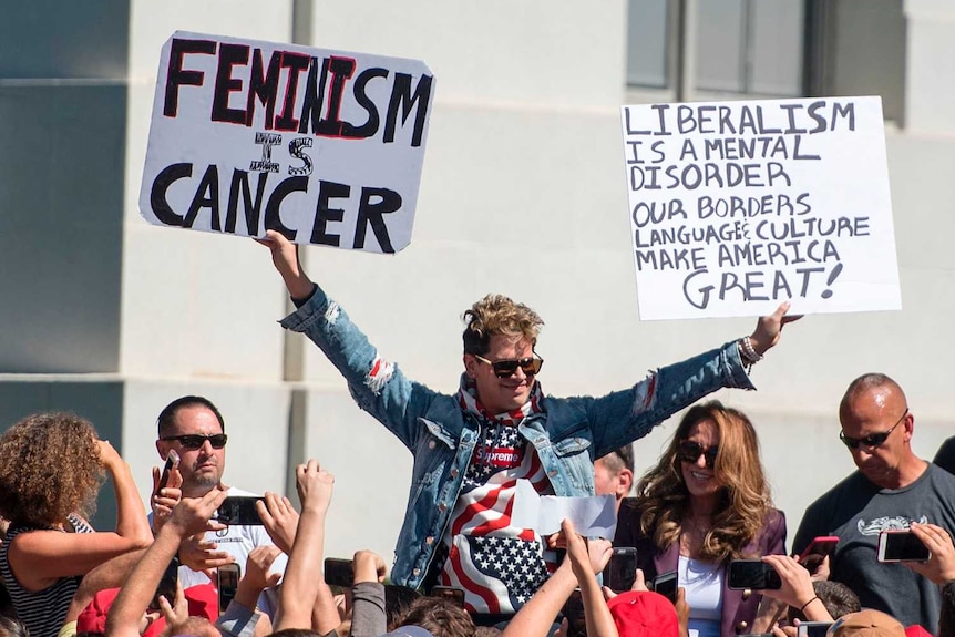 Milo Yiannopoulos holds up signs to a crowd of supporters on a university campus.