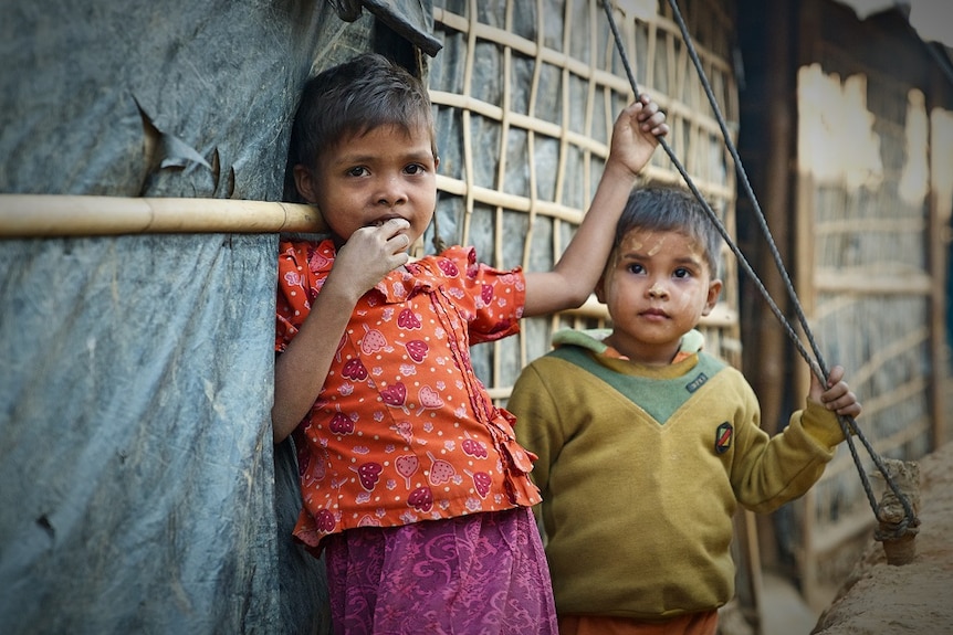 A young girl and a young boy stand outside a tent at a refugee camp.