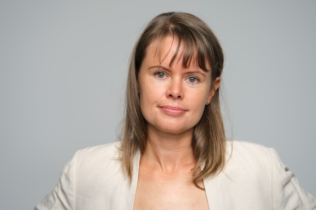 Bonney has snady brown hair, a fringe and blue eyes she is wearing a cream blazer and is standing behind a grey background