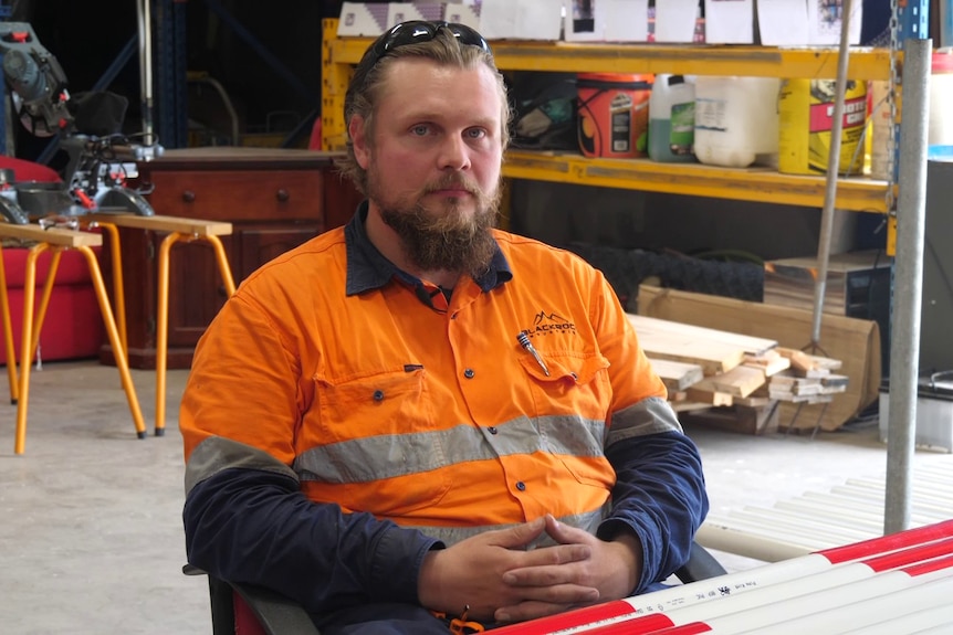 Dean, a young mine trainee wearing hi vis, sits in a workshop with his hands crossed over eachother looking at the came