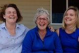 Three women sit on the top step of their house, all laughing.