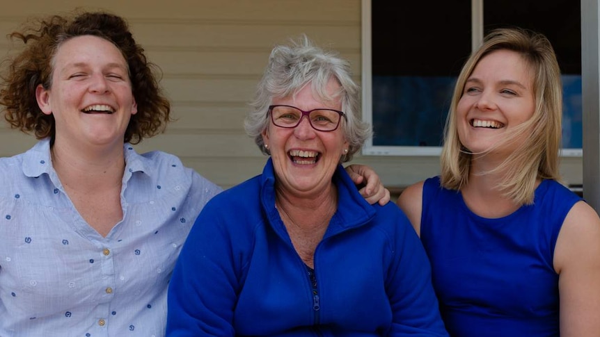 Three women sit on the top step of their house, all laughing.