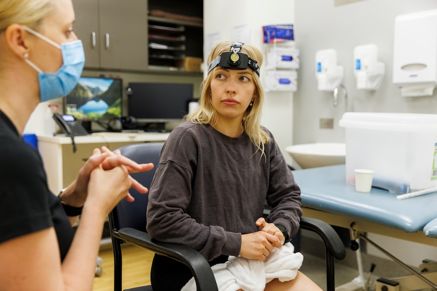 A woman with short blonde hair listens to a physiotherapist whilst wearing a laser head lamp commonly used in concussion rehab