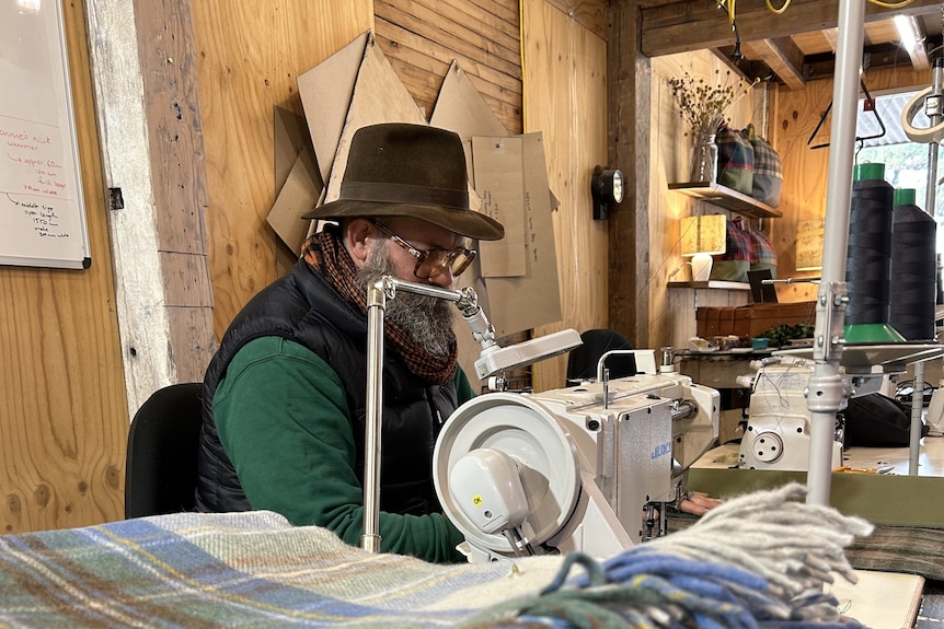 A bearded man wearing gloves and a hat sits at a sewing machine in a workshop.
