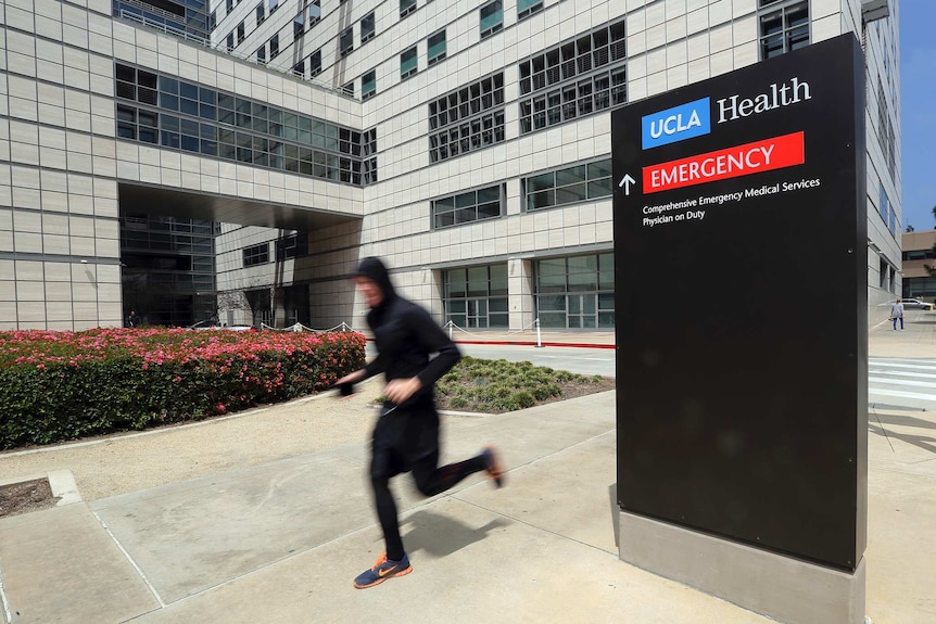 A runner walks past a sign that reads 'UCLA Health' in front of a large multi-storey medical complex.