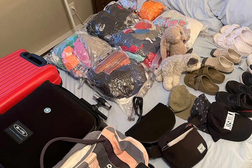 Bags of clothes and shoes line a bed next to a small suitcase.
