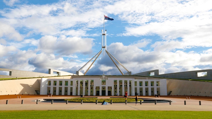 The front entrance of Parliament House, in the daytime, with flag raised and people walking around.