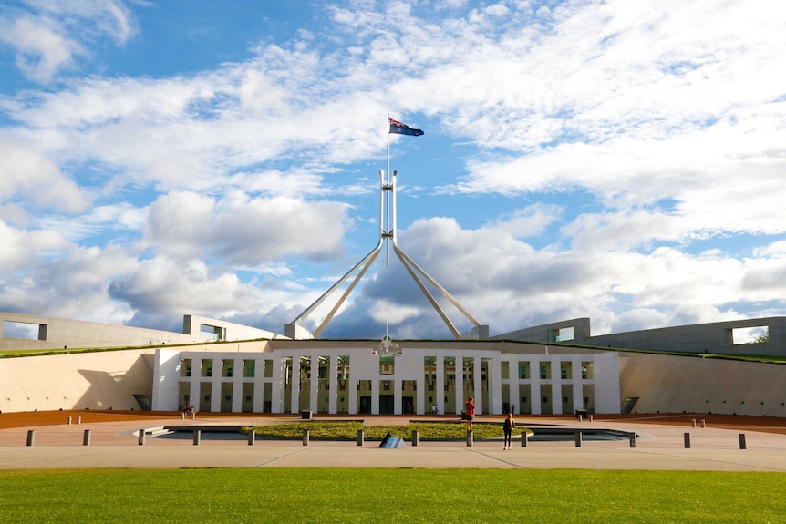 The front entrance of Parliament House, in the daytime, with flag raised and people walking around.