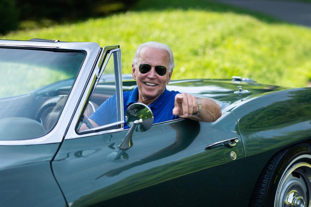 Joe Biden，身穿蓝色马球衫，戴着飞行员墨镜，坐在绿色敞篷车的方向盘后对着镜头。