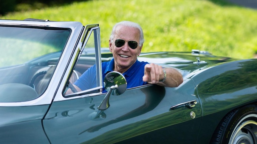 Joe Biden, wearing a blue polo shirt and aviator shades points at the camera from behind the wheel of a green convertible