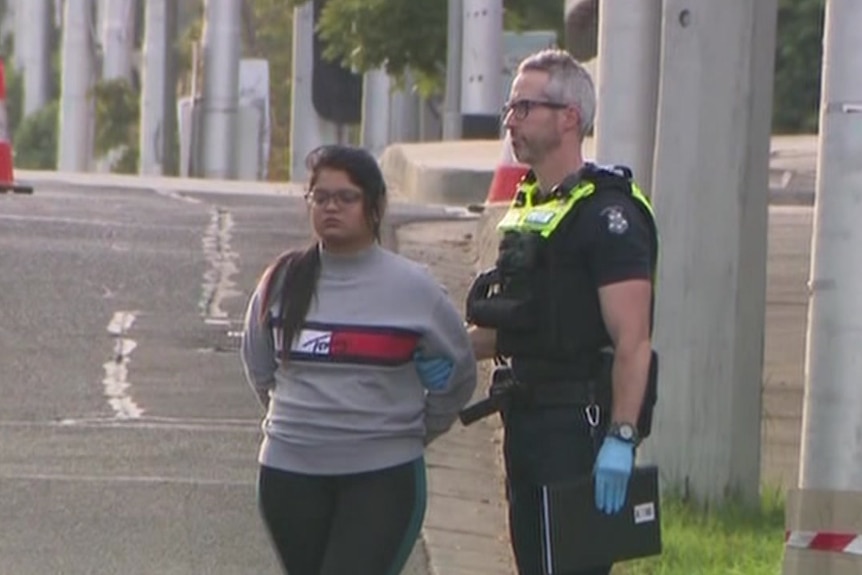 A woman wearing a grey jumper with her hands behind her back standing next to a police officer. 