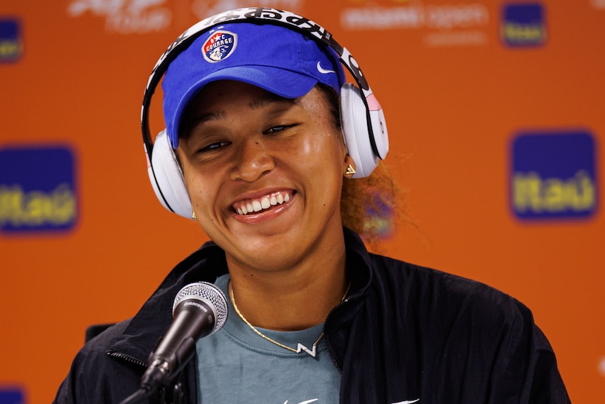 Naomi Osaka smiles during a press conference at the Miami Open wearing her headphones and hat