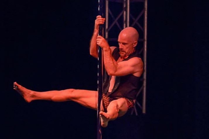 Man holds silver pole with both arms, while legs are spread wide and he is high off the ground