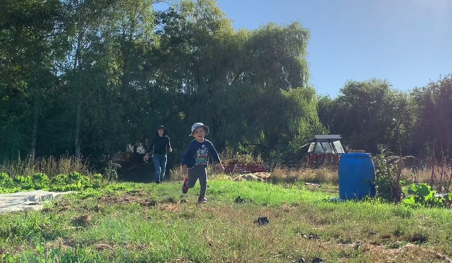 A little boy runs through a grassy area in front of big trees, as his mother follows from behind