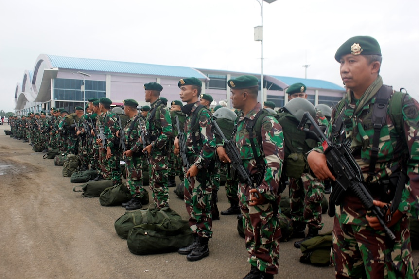 Indonesian soldiers in dark green camouflage and holding rifles line up with luggage on an airport tarmac.