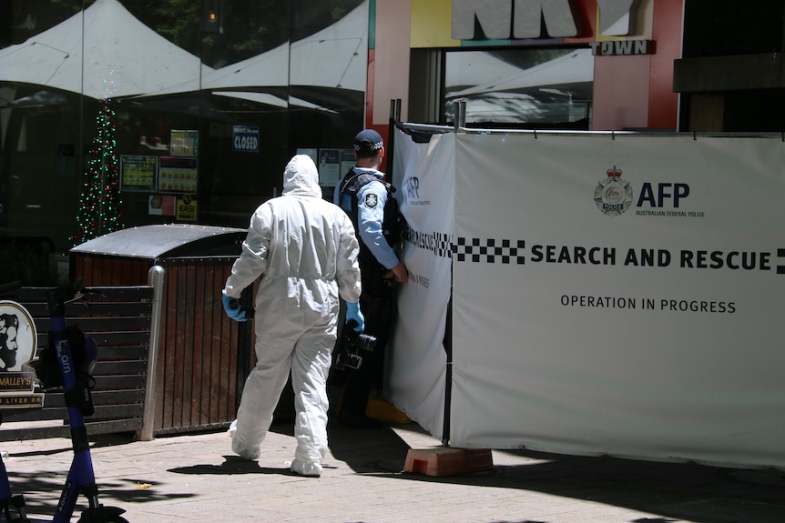 A police officer in uniform and a figure in a full-body white forensics suit stand by a police screen.
