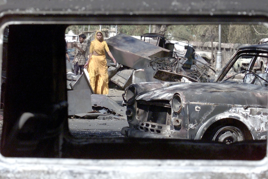 An Indian woman walks with her child between burned out cars in the street.