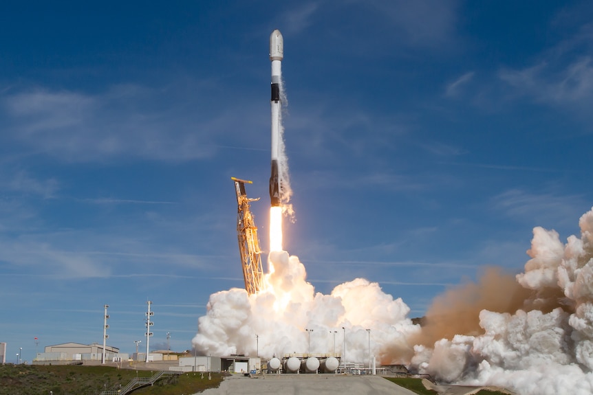 A wide shot of a rocket blasting off from a launch pad into a clear sky, with flames and clouds of smoke below it