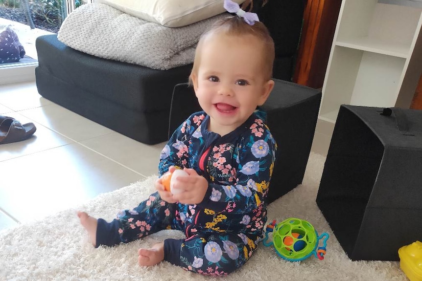 A baby wearing a hair tie smiles while playing with toys in a living room
