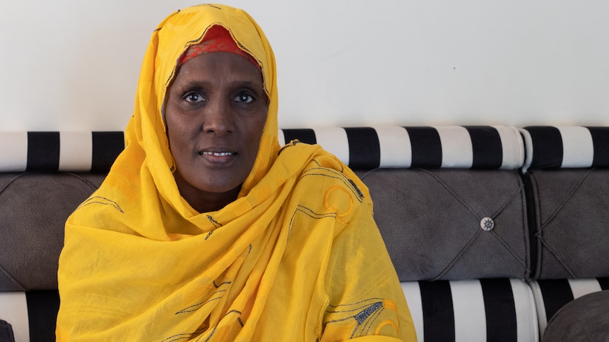 A woman in a yellow head scarf sits at home on a couch.