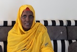 A woman in a yellow head scarf sits at home on a couch.