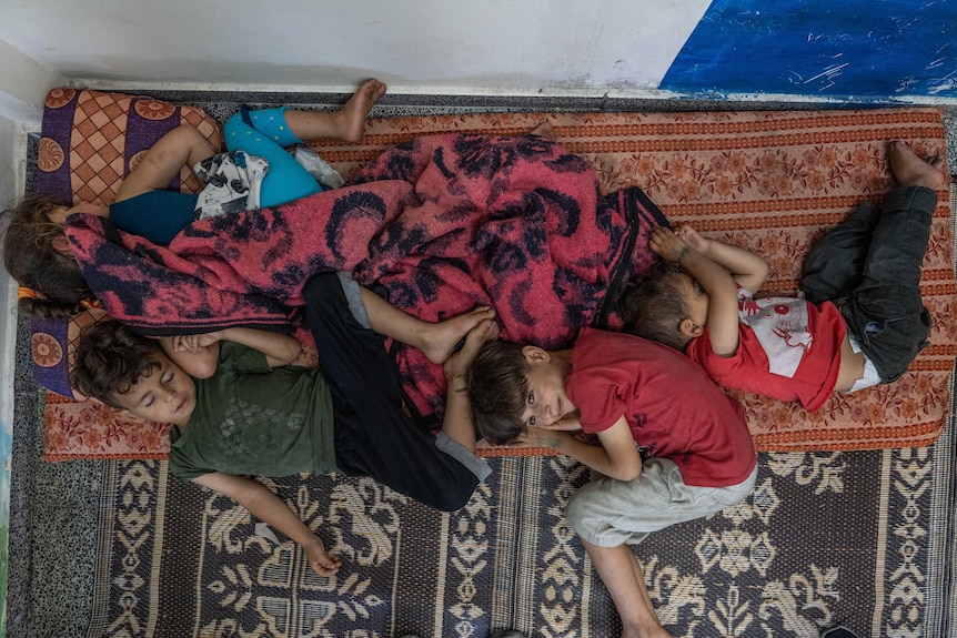 A group of young boys lie sleeping together on mats, with one awake and smiling 