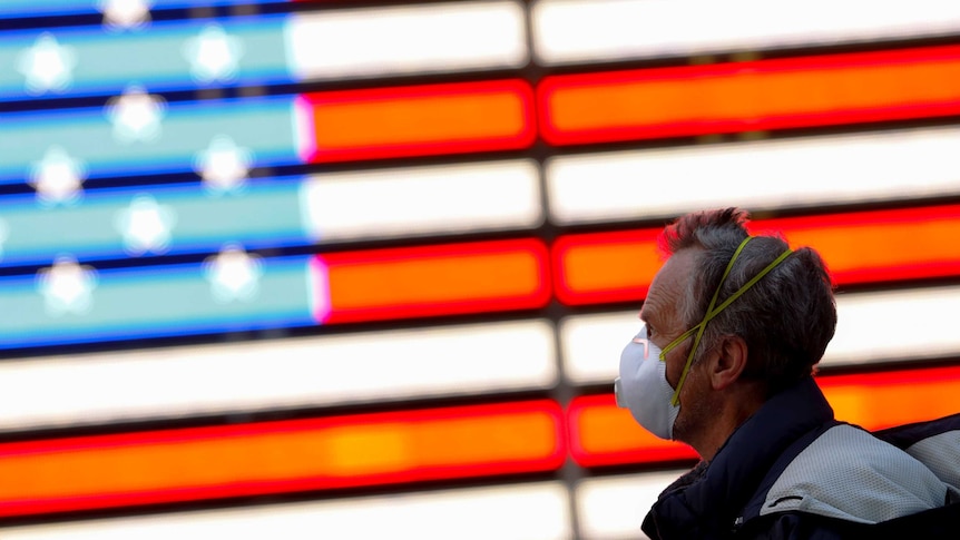 A man in a face mask walks past an American flag