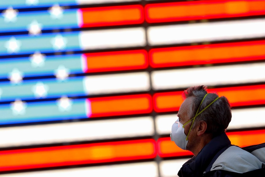 A man in a face mask walks past an American flag