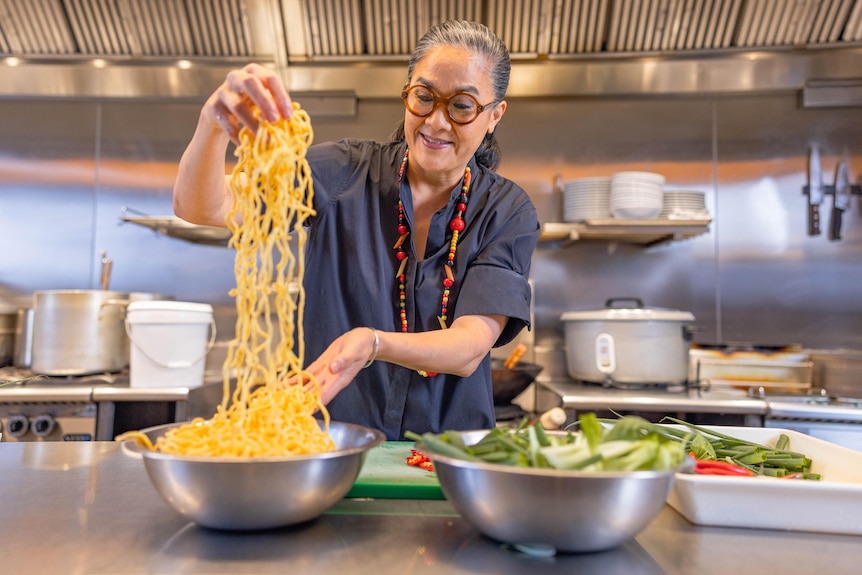Kylie Kwong holds noodles in her hand in a kitchen, with other ingredients in bowls on a kitchen bench.