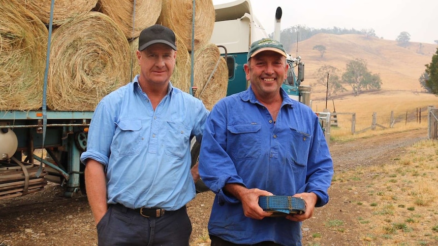 Two men wearing blue shirts and caps stand in front of a truck loaded with hay bales.