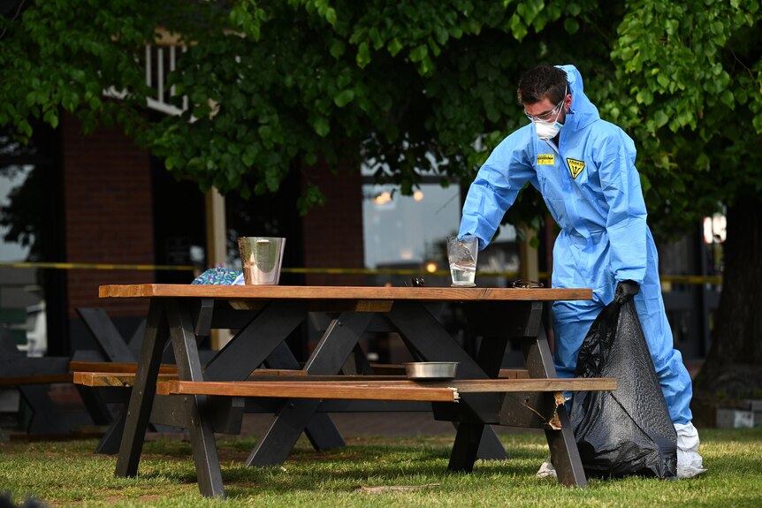 A person wearing blue PPE cleans up a glass on a picnic table.