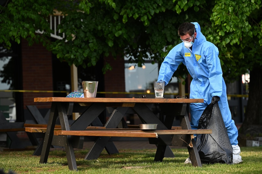 A person wearing blue PPE cleans up a glass on a picnic table.