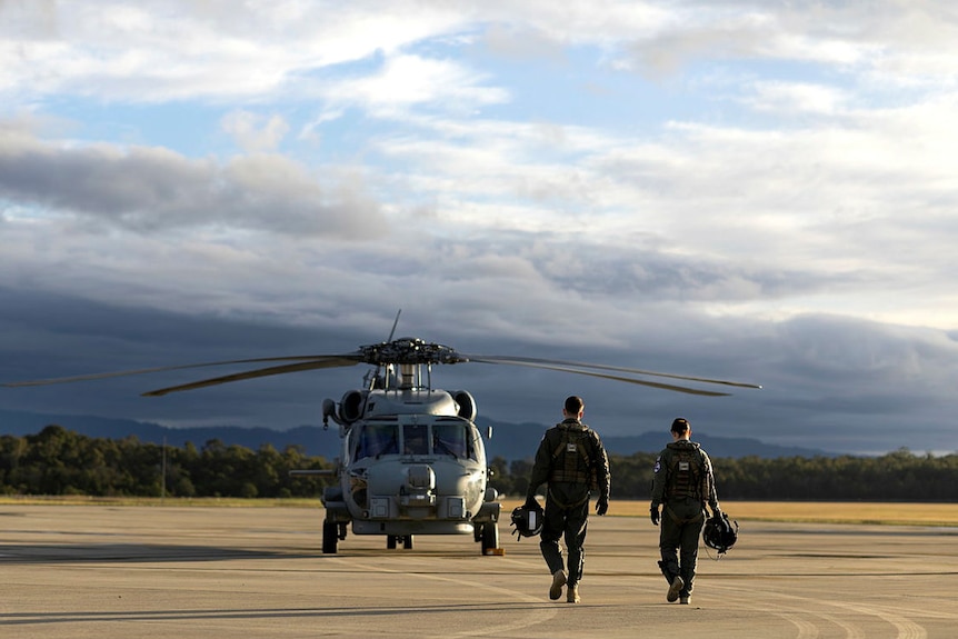 A helicopter sits on a tarmac as two men walk towards it