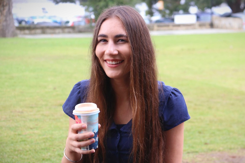 Woman sits on a park bench smiling and drinking a take-away coffee