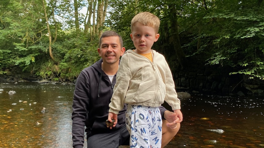 A young boy stands with his dad crouched next to him. There's a lake in the background.