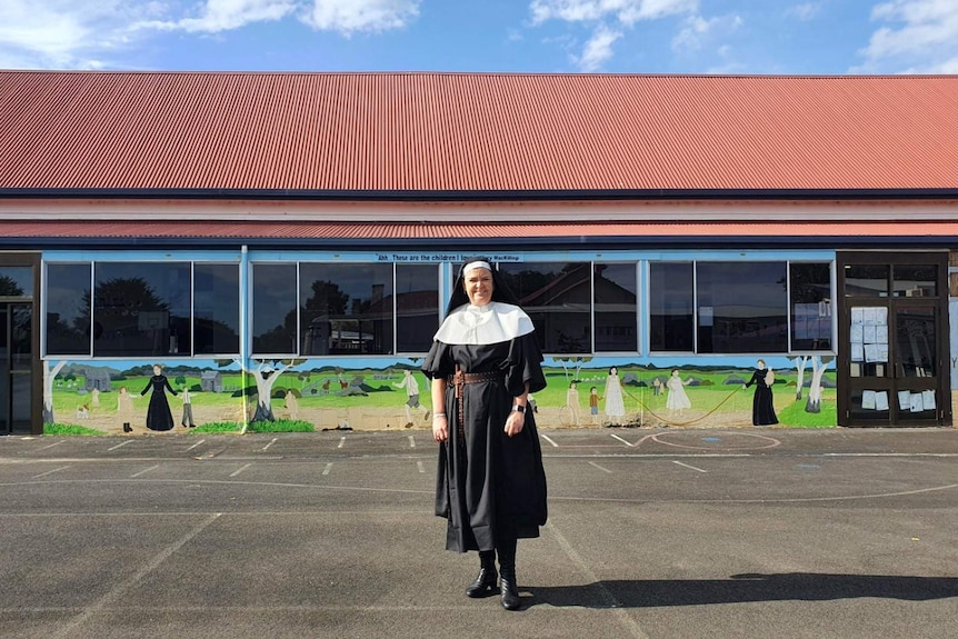 A woman dressed as a nun stands in a school yard with a colourful mural behind her.