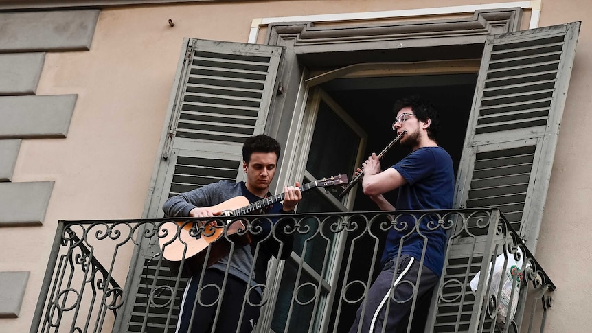 A man playing a flute and a man playing a guitar jam on a balcony.