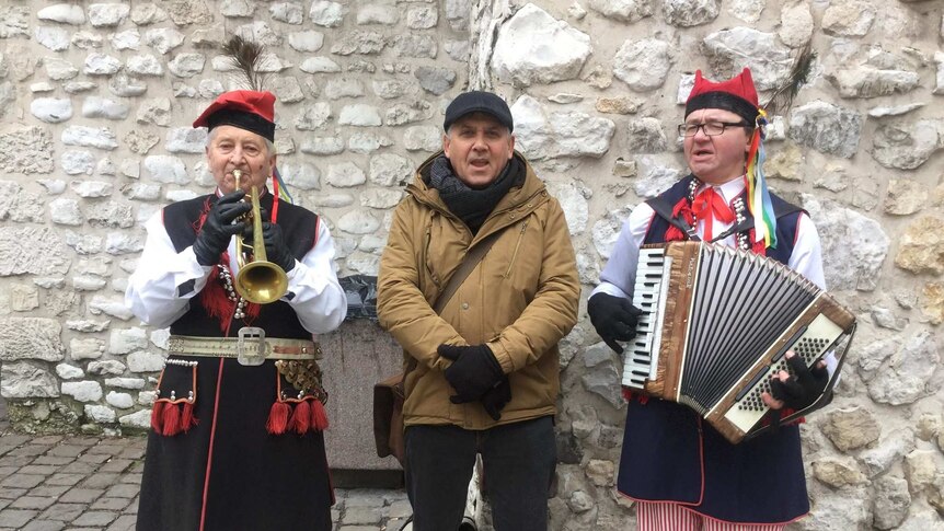 A man stands between two musicians in traditional Polish dress.