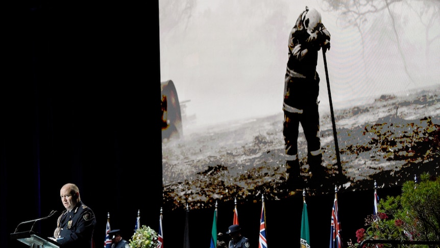 Commissioner Shane Fitzsimmons stands at a lectern at a funeral with a picture of a firefighter in the background