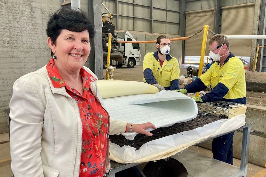 A middle-aged woman stand in the front as two male workers process an old mattress for recycling